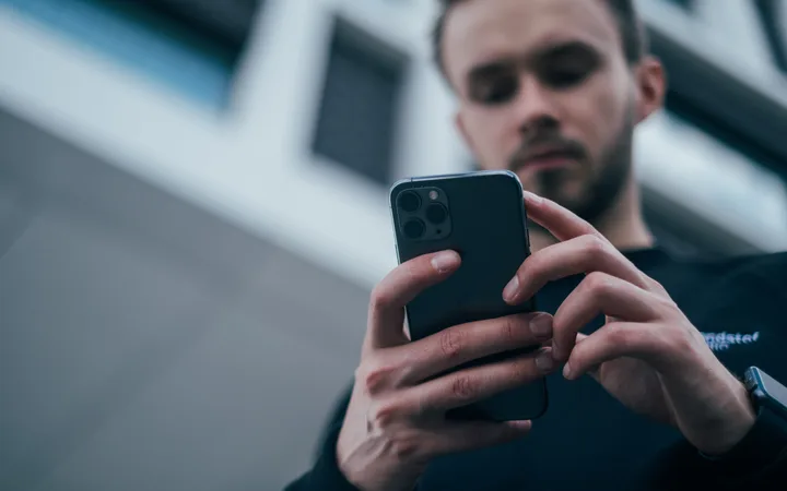 Person with dark hair and beard using a smartphone outdoors, with modern building architecture in the background.