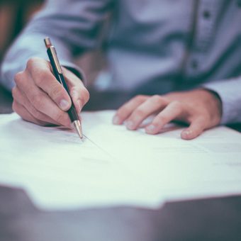 Person in a light blue shirt writing on a document with a black and gold pen, seated at a dark table, with focus on hands and pen.