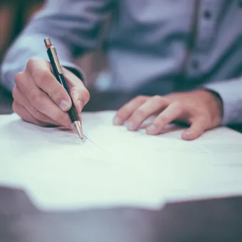 Person in a light blue shirt writing on a document with a black and gold pen, seated at a dark table, with focus on hands and pen.