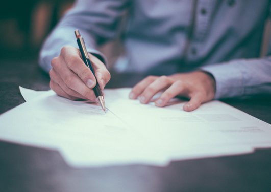 Person in a light blue shirt writing on a document with a black and gold pen, seated at a dark table, with focus on hands and pen.