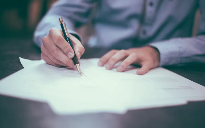Person in a light blue shirt writing on a document with a black and gold pen, seated at a dark table, with focus on hands and pen.