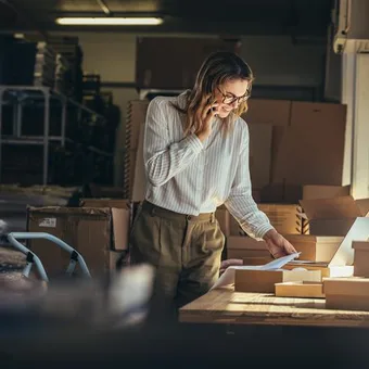 Woman in a warehouse reading a document, with packing boxes on a table, surrounded by shelves and natural light from a nearby window.