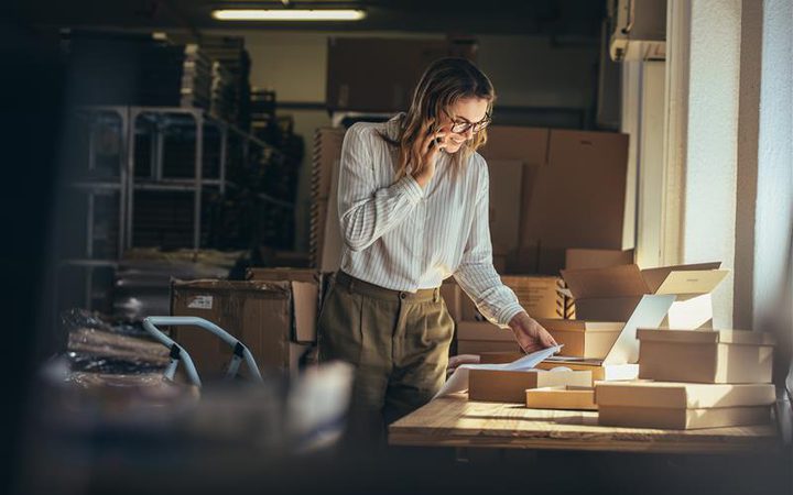 Woman in a warehouse reading a document, with packing boxes on a table, surrounded by shelves and natural light from a nearby window.