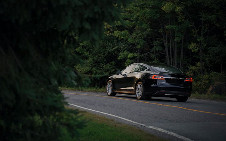 A black Tesla Model S drives along a wooded rural road, surrounded by dense green trees on a cloudy day.