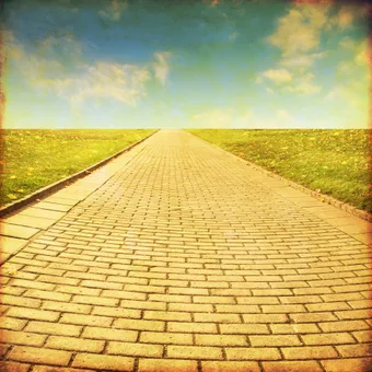 A long brick pathway stretches into the horizon, flanked by grassy fields under a partly cloudy sky, creating a sense of distance and openness.