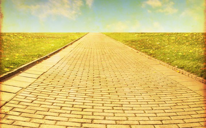 A long brick pathway stretches into the horizon, flanked by grassy fields under a partly cloudy sky, creating a sense of distance and openness.