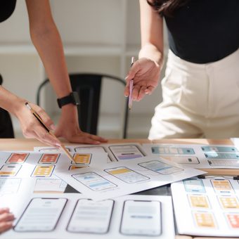 Two people stand around a table covered with mobile app wireframes and sketches, pointing with pencils.