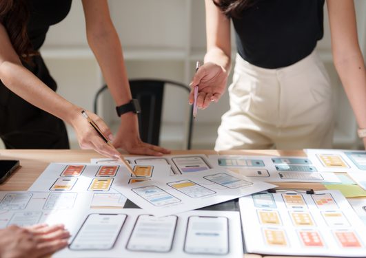 Two people stand over a table covered with colorful smartphone app wireframe sketches, pointing with pencils.