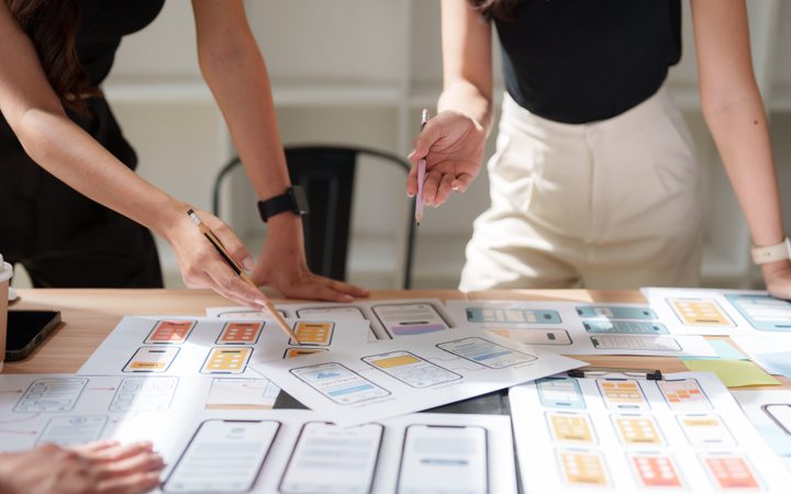 Two people stand around a table covered with mobile app wireframes and sketches, pointing with pencils.