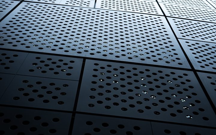 Close-up of a metallic perforated surface with circular holes arranged in grids, creating a pattern with reflections and shadows.