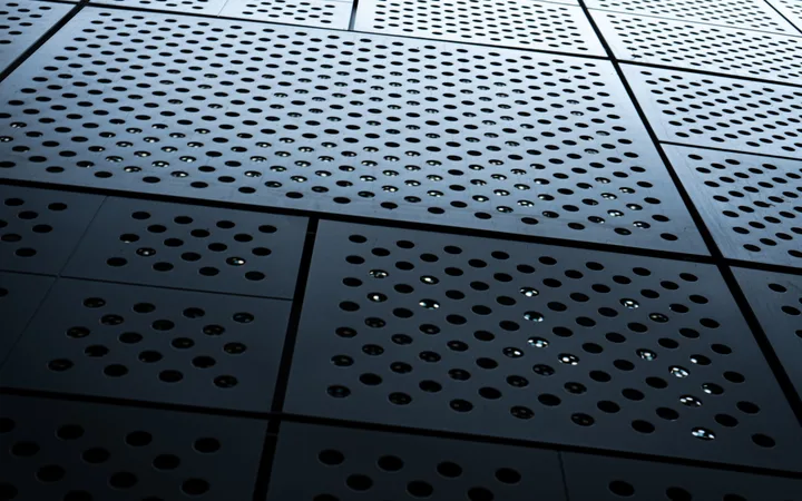 Close-up of a metallic perforated surface with circular holes arranged in grids, creating a pattern with reflections and shadows.