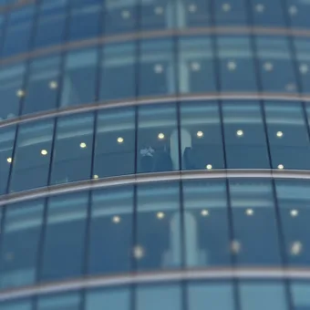 A person stands inside a glass-walled building, visible through the curved blue-tinted windows, with lights reflecting on the glass surface.