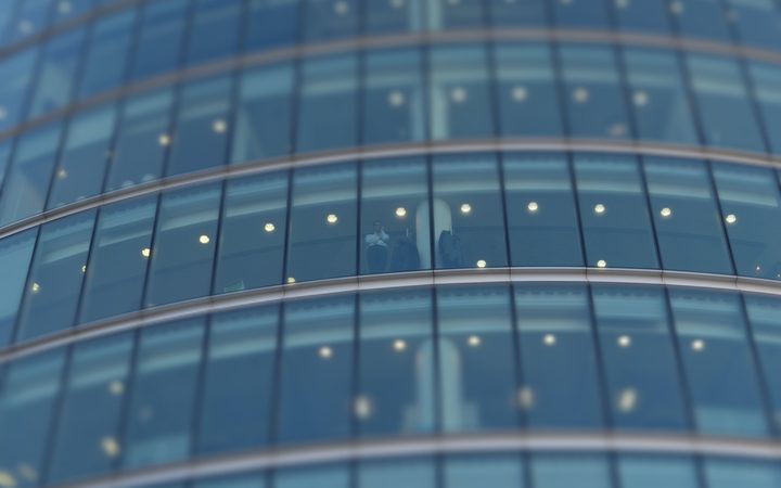 A person stands inside a glass-walled building, visible through the curved blue-tinted windows, with lights reflecting on the glass surface.