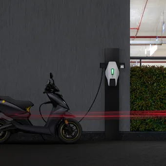 A black electric scooter parked at a charging station in a parking garage with a dark wall and some greenery in the background.