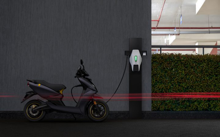 A black electric scooter parked at a charging station in a parking garage with a dark wall and some greenery in the background.