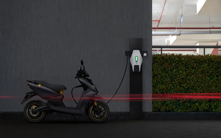 A black electric scooter parked at a charging station in a parking garage with a dark wall and some greenery in the background.