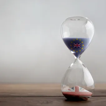 An hourglass with the European Union flag and UK flag symbols inside, resting on a wooden surface with a plain light background.