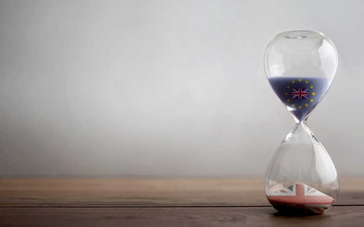 An hourglass with the European Union flag and UK flag symbols inside, resting on a wooden surface with a plain light background.