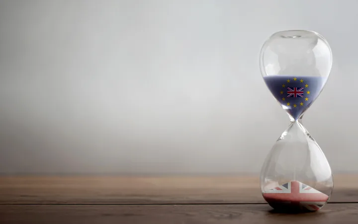 An hourglass with the EU flag and the UK flag reflected inside, placed on a wooden surface against a plain, light grey background.