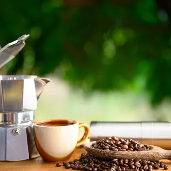 Coffee-making setup with a moka pot, a beige mug filled with coffee, a wooden spoon with coffee beans, and a metal travel mug on a wooden surface.