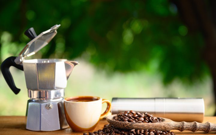 Coffee maker, mug, coffee beans, and a spice grinder on a wooden surface outdoors with blurred green foliage in the background.
