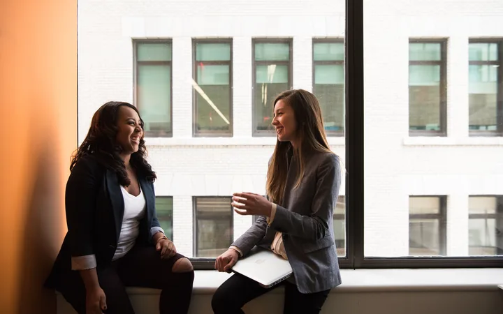 Two people are smiling and talking by a large window in an office building, one holding a laptop while sitting on the window ledge, the other standing.