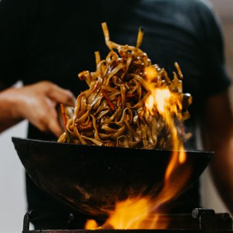 Person in a black shirt tossing a mound of glossy stir-fried noodles in a wok over an open flame.