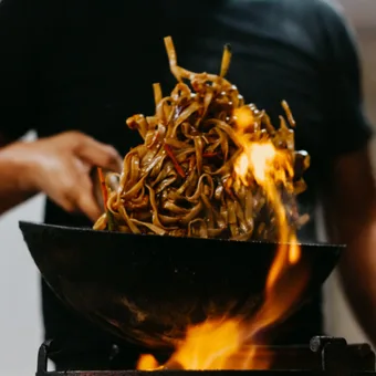 Person in a black shirt tossing a mound of glossy stir-fried noodles in a wok over an open flame.