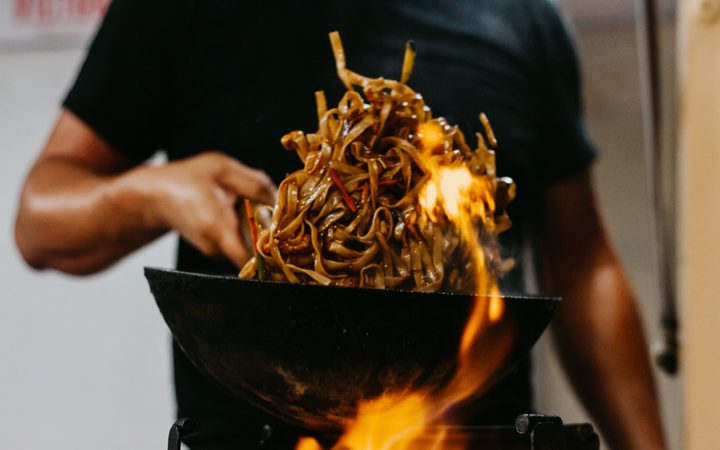 Person in a black shirt tossing a mound of glossy stir-fried noodles in a wok over an open flame.