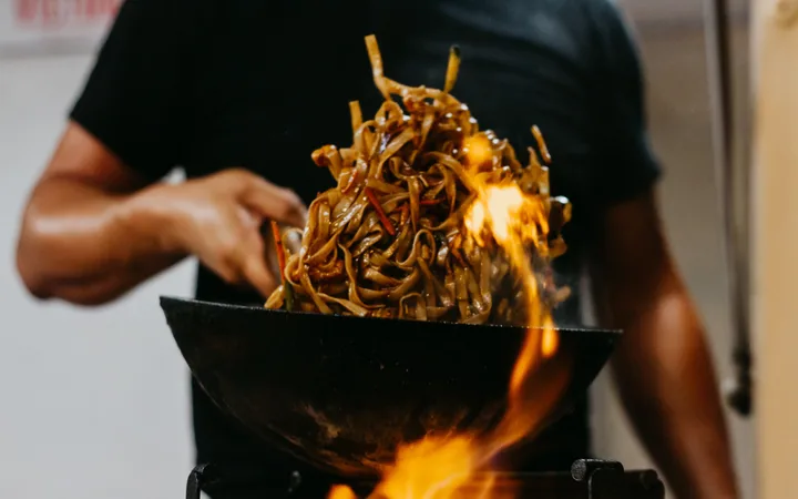 Person in a black shirt tossing a mound of glossy stir-fried noodles in a wok over an open flame.