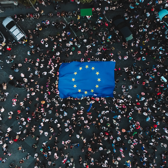 Aerial view of a crowded street with a large European Union flag laid on the ground, surrounded by people and parked cars.