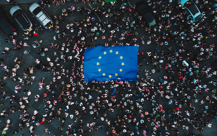 Aerial view of a crowded street with a large European Union flag laid on the ground, surrounded by people and parked cars.