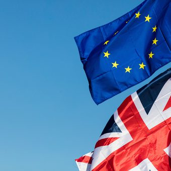 Two flags flutter in a clear blue sky: the European Union flag with yellow stars and the United Kingdom flag beneath it.
