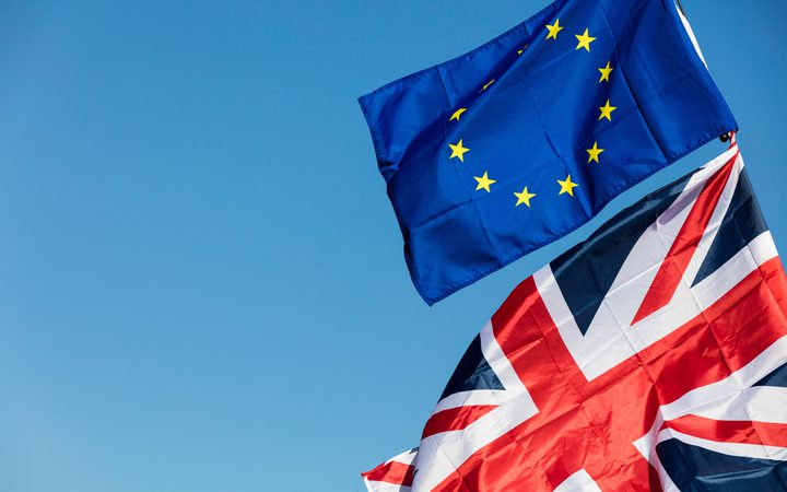 Two flags flutter in a clear blue sky: the European Union flag with yellow stars and the United Kingdom flag beneath it.