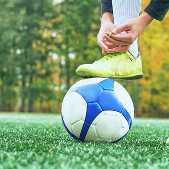 A person wearing yellow sports shoes and black socks is balancing a blue and white soccer ball on their foot on a green field with a goal in the background.