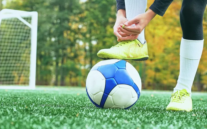A person wearing yellow sports shoes and black socks is balancing a blue and white soccer ball on their foot on a green field with a goal in the background.
