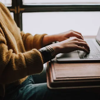 Person in a tan sweater typing on a laptop at a wooden table near a window.