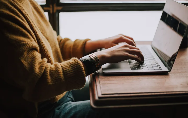 Person in a mustard sweater working on a laptop at a wooden desk by a window.
