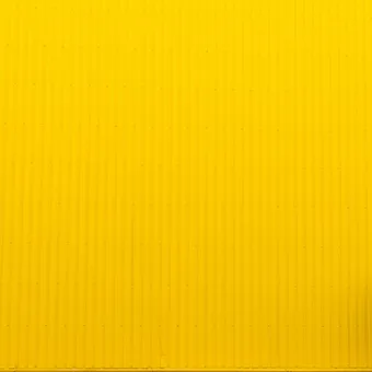 A woman walks past a large, yellow corrugated metal wall, positioned on the right side of the image, with a bright and minimalist composition.