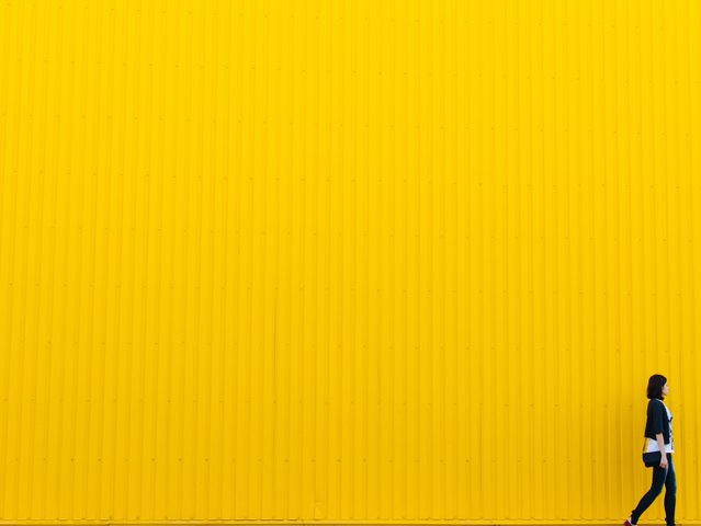 Femme marchant devant un grand mur jaune en métal, vue de profil, portant un sac à bandoulière et un manteau noir.