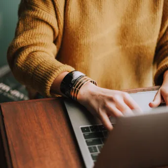 Person wearing a mustard sweater working on a laptop at a wooden table, with a book partially visible in the background.