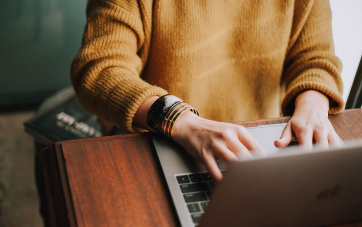 Person wearing a mustard sweater working on a laptop at a wooden table, with a book partially visible in the background.