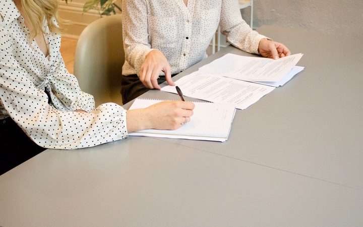 Two women in polka dot blouses review documents and take notes at a desk in an office setting.