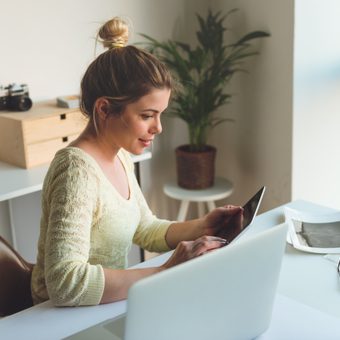 Person with a light bun wearing a cream lace top sits at a white desk, using a tablet beside a laptop, in a bright home office with plants and a window.