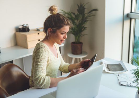 Person with a light bun wearing a cream lace top sits at a white desk, using a tablet beside a laptop, in a bright home office with plants and a window.
