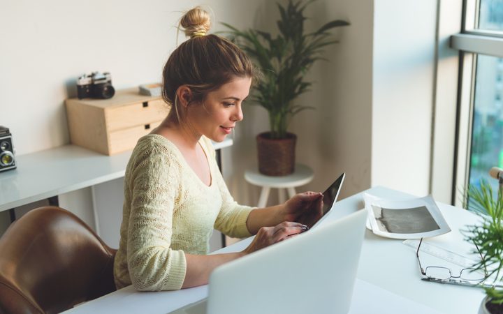 Person with blond hair in a bun, wearing a pale yellow knit sweater, sits at a bright desk using a tablet beside a laptop, with plants and a window nearby.