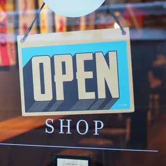 Glass door with a hanging "OPEN" sign and "SHOP" text below, showing reflections of colorful storefronts and lights inside the store.