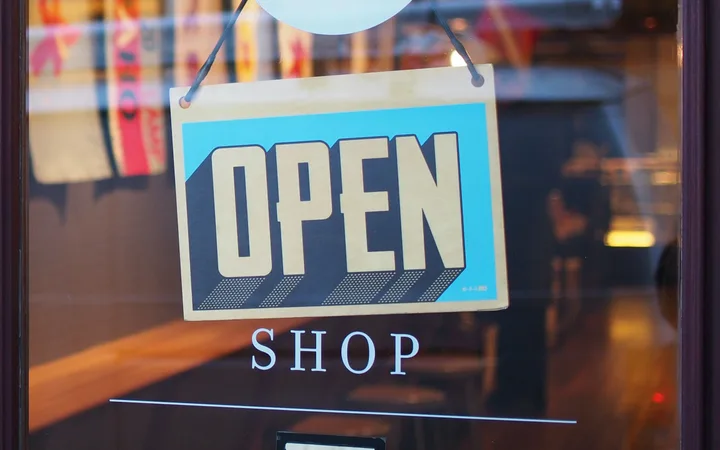 Glass door with a hanging "OPEN" sign and "SHOP" text below, showing reflections of colorful storefronts and lights inside the store.