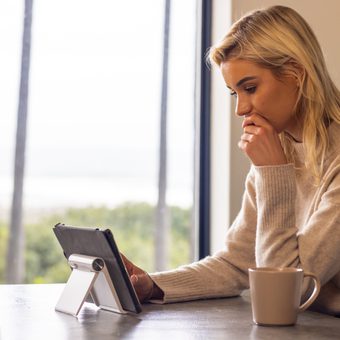 Person with blond hair in a beige sweater sits at a table by a window, looking at a tablet on a stand, with a mug nearby.