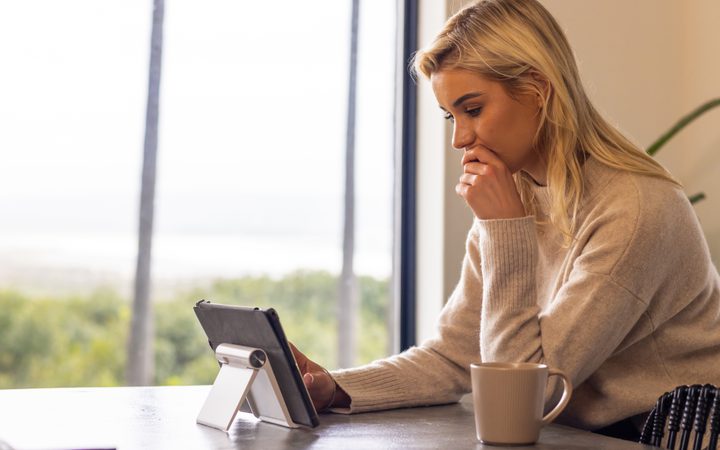 Person with blond hair in a beige sweater sits at a table by a window, looking at a tablet on a stand, with a mug nearby.